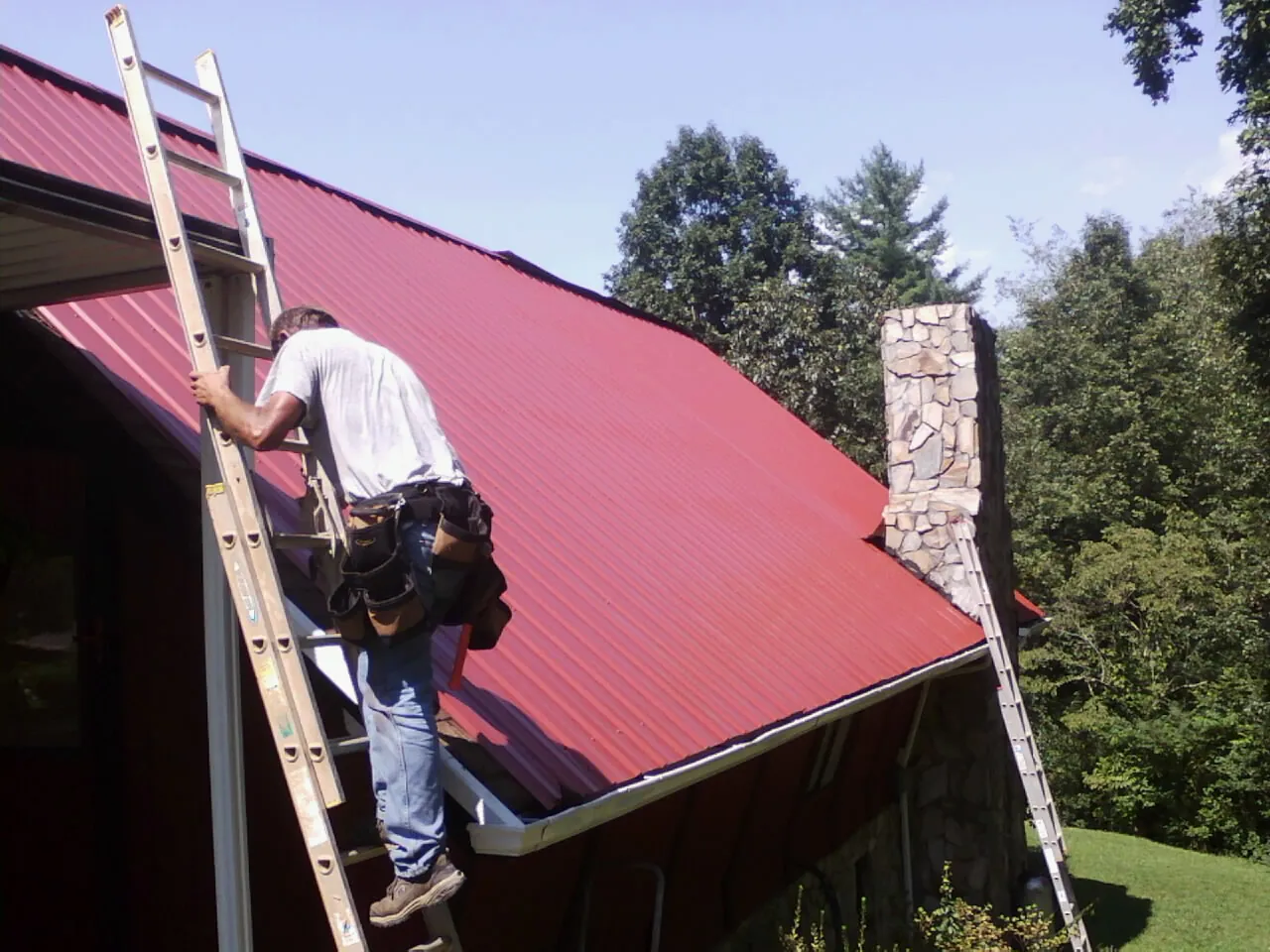 Roofer on a ladder beside a red metal roof and stone chimney — Soffit & Fascia Repair by Roofing Brothers in Charlemont